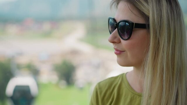 Positive long haired woman with black sunglasses looks with interest at moving cabin of poma lift at mountain station closeup slow motion