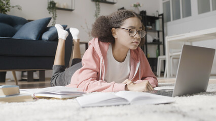 Curly-haired teenage girl listening to online lessons lying in front of laptop