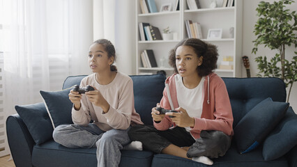 Excited friends playing video game with joysticks at home, sitting on the sofa