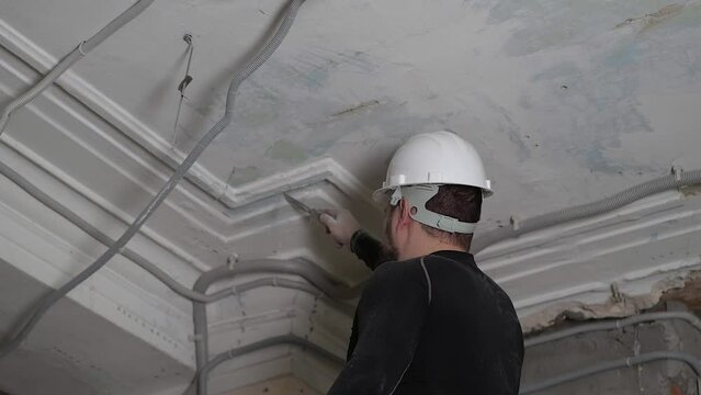 Fat bearded adult man in protective helmet and black tight fitting clothing remove construction whitewash vintage high ceiling. Overhaul repair with your own hands concept. 4k slow motion footage 
