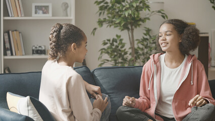 Two black teen girls talking on couch, friends having fun together at home