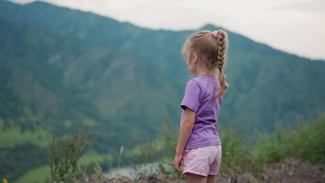 Cute little girl with braided hair looks at old distant mountain foggy silhouettes and scratches leg at highland backside view slow motion