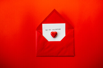 Red envelope with a note "Be my valentine" on a red background with chocolate candies in the shape of a heart in red foil.