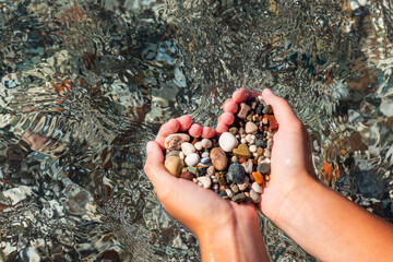 Hands full of colorful pebbles forming heart shape on the sea waves background. Holiday, rest, vacation at the seaside