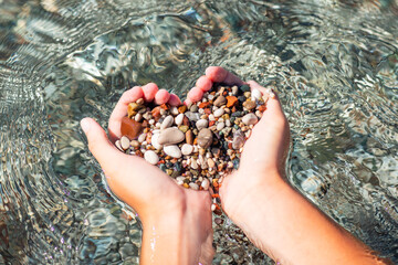 Hands full of colorful pebbles forming heart shape on the sea waves background. Holiday, rest, vacation at the seaside