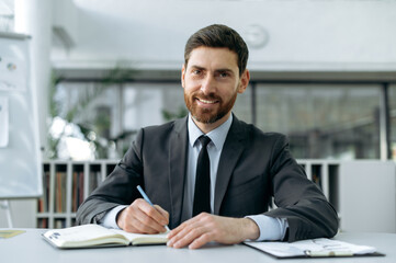Obraz premium Confident intelligent caucasian businessman, sit in modern office, listening lecture, webinar via video conference, taking notes in a notebook, communicates with business partners, conducts briefing