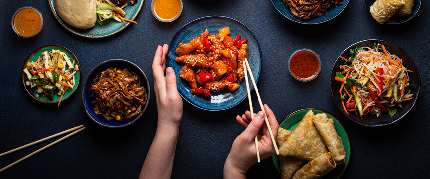 Set of Chinese dishes on table, female hands holding chopsticks: sweet and sour chicken, fried spring rolls, noodles, rice, steamed buns with bbq glazed pork, Asian style banquet or buffet, top view 
 - Powered by Adobe