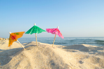 Three small paper cocktail umbrellas stand in sand on sandy beach close-up. Small paper umbrellas on sandy shore near sea and sea waves on sunny summer day. Leisure, Vacation, Travel, Tourism Concept