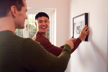 Same Sex Male Couple Hanging Picture On Wall At Home Together
