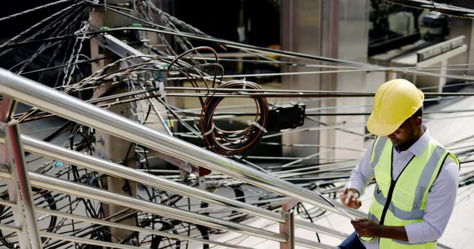 Electrical engineer checking electrical wires Internet cables on electric poles in city. African American technician check data. Repairing from tablet with expertise. supervise construction site