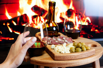Female hand hold wine glass against wine table with appetizers for wine and electric fireplace. Manicure with red hearts. Romantic dinner at home concept.