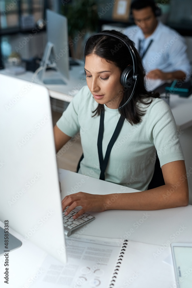 Poster Dialled in and getting it done. Shot of a young woman using a headset and computer in a modern office.
