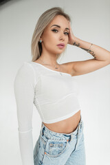 Naklejka premium Fashion cute girl with blonde hairstyle in stylish white t-shirt and blue jeans stands and looks at the camera on a white background in studio