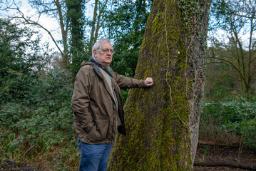 Portrait of active senior man walking outdoors in the woods, resting on a tree