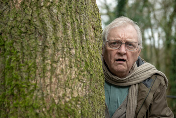 Portrait of shocked senior man peeking from behind a large tree in the woods