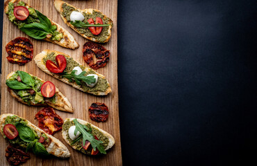 Set of bruschettas with pesto, guacamole, grilled tomatoes and arugula on wooden board with copy space. Italian tosted bread with vegetables, mozzarella cheese, basil and traditional sauces