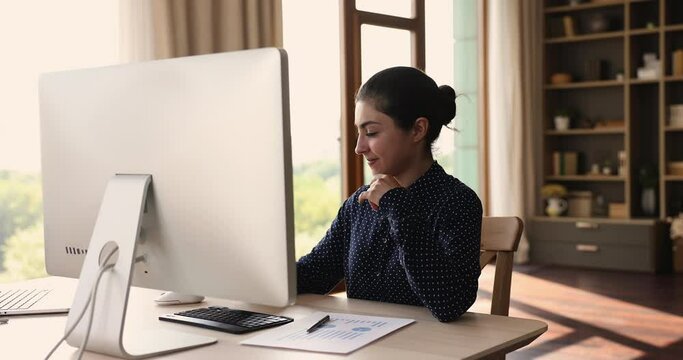 Attractive young Indian ethnicity woman sit at workplace desk looks at pc screen do stocks data analysis, analyze sales statistic feels satisfied by results, prepare financial report, trading concept