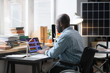 Rear view of African architect sitting in wheelchair at his workplace and typing on laptop, he developing new project online