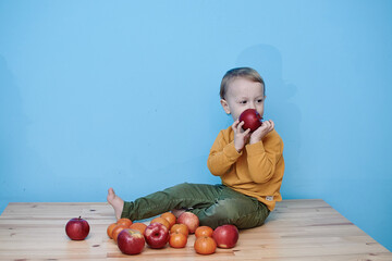 Fototapeta premium a little boy of 2 years old sits on a table against a blue wall and holds a red apple. there are a lot of red apples and a lot of tangerines around. child in green pants and yellow sweater