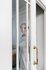 A woman in a white airy poses in front of a glass door. A bride on her wedding day.