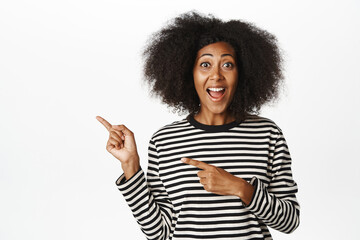 Surprised happy african woman, pointing fingers left, showing copy space, empty place for announcement, store promo deal, standing over white background