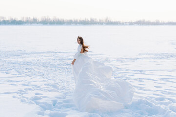 Fototapeta na wymiar Incredible stunning girl in a white airy dress. In the background is a winter landscape of a frozen river covered in ice. Artistic photo