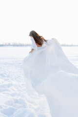 Fototapeta na wymiar Incredible stunning girl in a white airy dress. In the background is a winter landscape of a frozen river covered in ice. Artistic photo