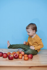 Fototapeta premium a little boy of 2 years old sits on a table against a blue wall and holds a red apple. there are a lot of red apples and a lot of tangerines around. child in green pants and yellow sweater