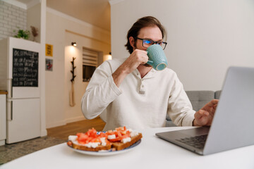 Ginger bristle man in eyeglasses working with laptop while having dinner