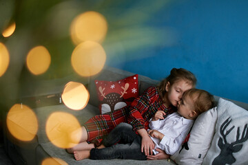 a girl in a red dress 8 years old and a boy in a white shirt 2 years old. brother and sister sit on the couch near the Christmas tree. they are friends and love each other
