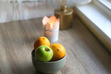 Bowl of lemons and apples, candle holder with lit candle and vase with gypsophila flowers on the table. Bokeh lights in the background. Selective focus.