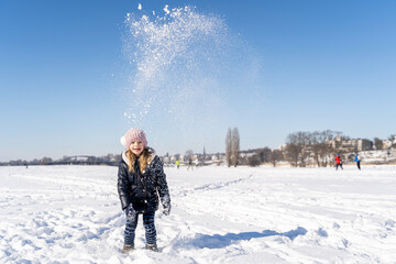 Young caucasian smiling preschool girl in warm clothes throws snow up playing and having fun on sunny winter weather outside on snow covered field in city park. Christmas holidays, childhood, carefree