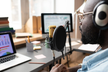Close-up of African man in headphones speaking to the microphone while sitting at the table and using laptop during his podcast