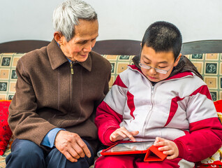 Senior Man and Young Boy Reading at Home. A teenage boy is looking down at a tablet computer, finger touching the screen. The grandfather is watching..