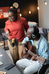 Fototapeta na wymiar African young woman singing song while her friend playing guitar sitting at the table in front of the microphone during online broadcasting