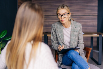 Female therapist in glasses listens attentively to the patient and writes down the diagnosis. - obrazy, fototapety, plakaty