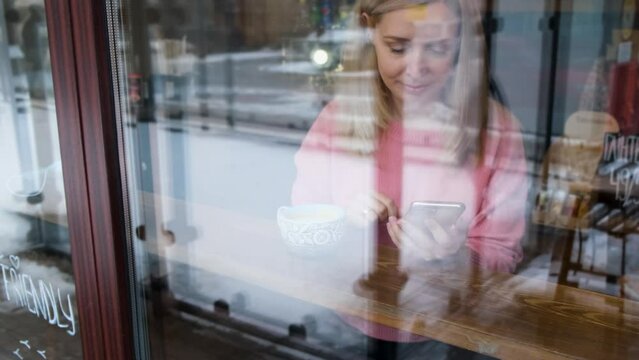 a blonde girl sits inside a cafe behind glass drinking coffee and scrolling through the tape in the phone