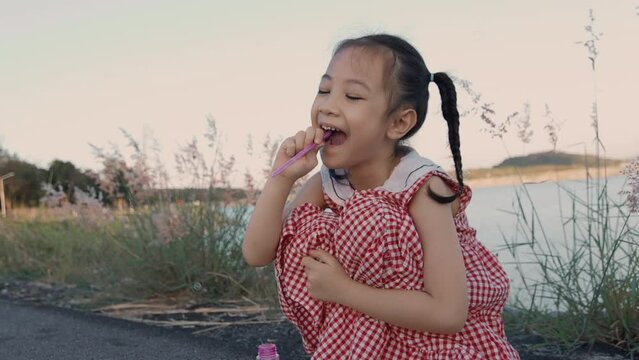 Face focus with copy space of asian girl is blowing soap-bubble in the park with beautiful sky and lake during sun set. It shows leisure time outdoor activity with enjoyment and cheerful expression.