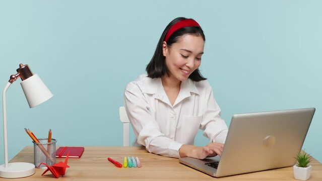 Fun young successful employee business woman of Asian ethnicity 20s in casual shirt sit work at office desk typing on pc laptop computer isolated on plain pastel light blue background studio portrait