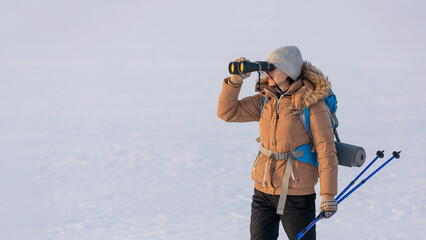 Woman in down jacket with a backpack and ski poles in hand looks through binoculars standing on a snowy plain in winter, selected focus. © junky_jess