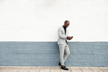 Full body smiling african american businessman leaning against wall on street with cellphone
