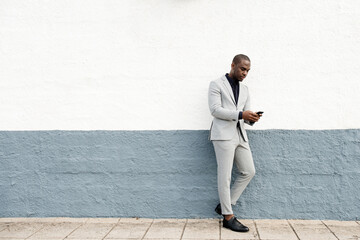 Full body serious african american businessman leaning against wall by street with cellphone