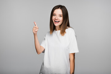 happy brunette woman showing thumb up while looking at camera isolated on grey