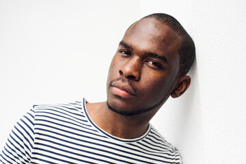 Close up handsome black guy in striped shirt leaning against white wall staring