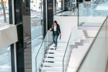 Businesswoman working with a digital tablet while drinking coffee going down the office stairs.