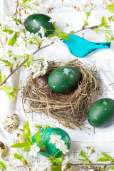 Fototapeta na wymiar Easter eggs colored with nettles in green color on table in nest and around it on white tablecloth in green wooden bird and flowering cherry branches background, Easter holiday table decoration
