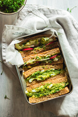 Healthy, natural summer food. Serving of sandwiches with whole grain bread, avocado, eggs, radish, cucumber in a zero waste metal lunchbox on a napkin on a white wooden background