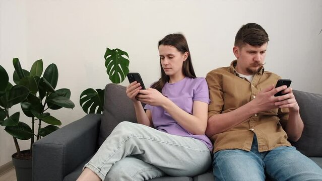 Young couple holding smartphones, looking at phone, playing mobile game, using mobile cell app together sitting on cozy grey sofa in living room at home, betting or bidding in auction application