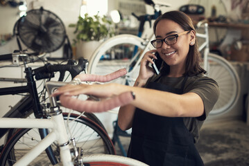 Ive got your bike ready for pickup. Shot of an attractive young woman standing alone in her shop and repairing a bicycle wheel while using her cellphone.