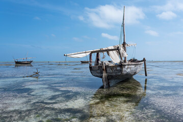  Traditional wooden boat with lowered sail at low tide with blue cloudy sky in Zanzibar, Tanzania
